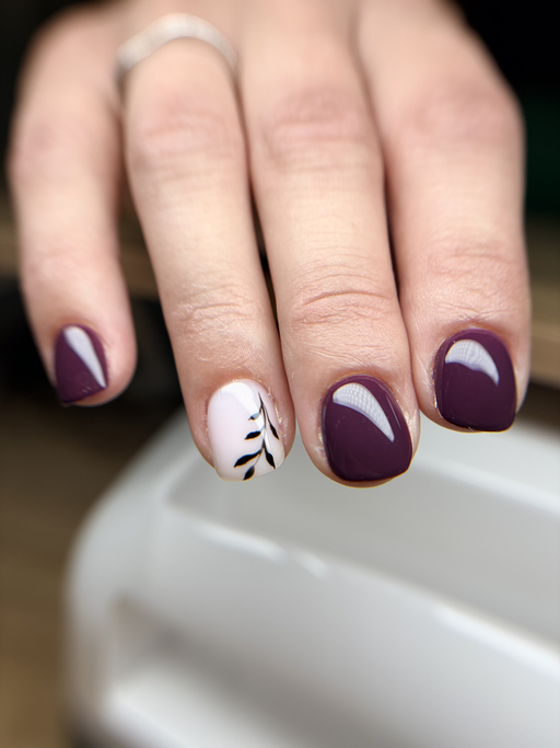 Ongles carrés courts, prune brillant, un ongle rose pâle orné d’une fine branche de feuillage noir.
