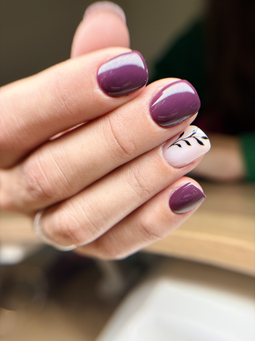 Ongles carrés courts, prune brillant, un ongle rose pâle orné d’une fine branche de feuillage noir, main levée.