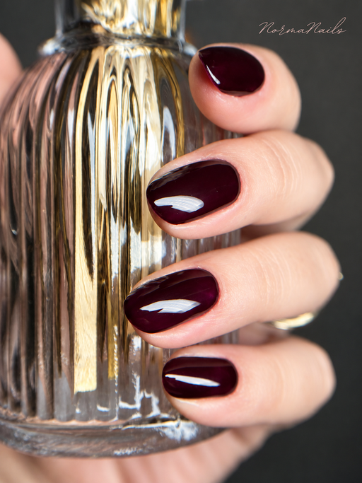 Ongles courts arrondis, bordeaux profond uni à la finition laquée, posés sur un flacon de parfum doré.
