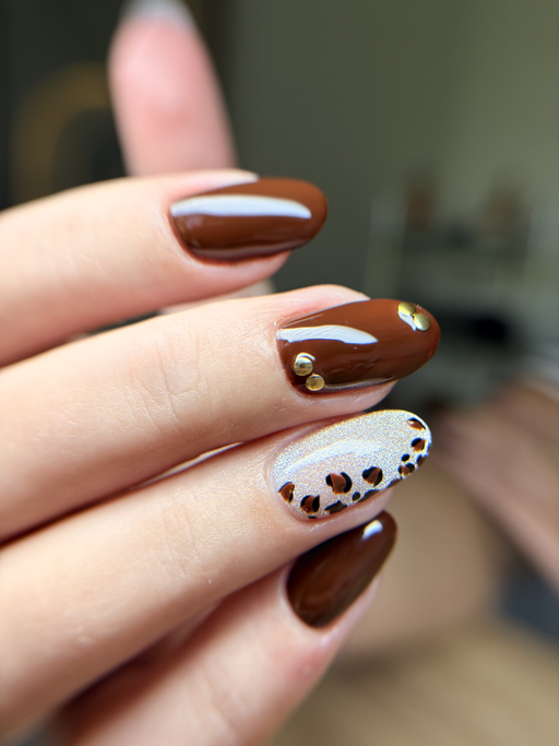Ongles amande de longueur moyenne, chocolat brillant, un ongle beige à motif léopard et un ongle orné de deux clous dorés, main vue de profil.